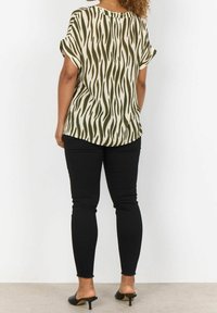 Blusa a maniche corte di colore crema con motivo ondulato verde, abbinata a jeans neri slim-fit e mule nere. Presenta un orlo arrotondato e una vestibilità rilassata.