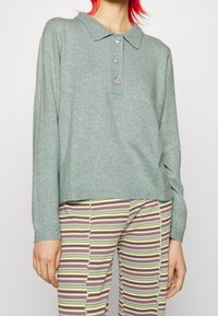 Pull en maille vert menthe avec col, doté de trois boutons en forme de bijoux et de poignets côtelés, associé à un pantalon rayé multicolore.