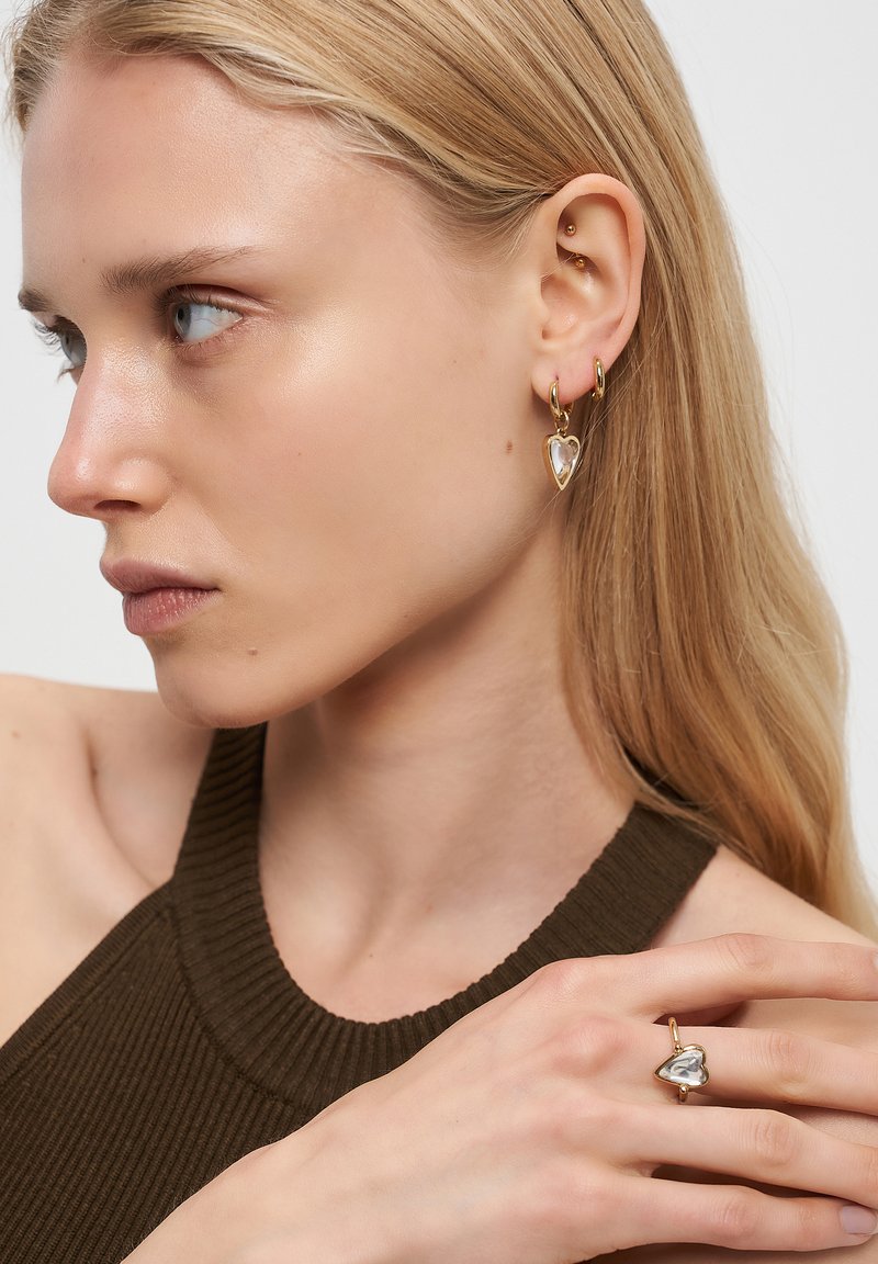 Blond kvinna som bär guldörhängen och matchande ring med klara hjärtformade stenar, klädd i en ribbad mörkbrun ärmlös topp.