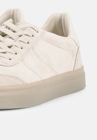 Ανοιχτό μπεζ sneaker από σουέτ με επίπεδα κορδόνια, λείο καουτσούκ πέλμα και διακριτικές ραφές σε τόνους στο επάνω μέρος. Μινιμαλιστικός σχεδιασμός χωρίς σχέδια.