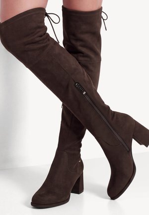 Mujer con chaleco de piel marrón oscuro sin mangas, jersey de cuello alto blanco, falda marrón oscura y botas altas hasta el muslo, sosteniendo un bolso beige texturizado.