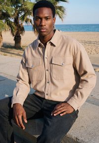Jeune homme aux cheveux courts portant une chemise beige à boutons et un pantalon noir, assis à l'extérieur près d'une plage de sable avec des palmiers et l'océan en arrière-plan.