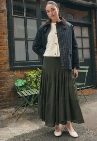 Plaid navy jacket with brown collar, off-white fleece top, and dark green tiered maxi skirt. Beige slingback shoes. Brick and glass background.
