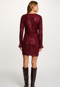 Lyhyt burgundinpunainen paljettipuku, jossa pitkät hihat ja kellomaiset kalvosimet. Pukeutunut polven korkeisiin ruskeisiin nahkasaappaisiin. Sileä tekstuuri.