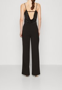 Zwarte mouwloze top met een diepe ruglijn, versierd met zilveren studs, gecombineerd met hoge zwarte broek met wijde pijpen en hakken.