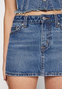 Denim mini kjol i blekt blått, med fransad nederkant, femfickorsdesign och kontrasterande orange sömmar samt metallknapp stängning.