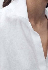 Chemise en lin blanc avec un col classique, une texture lisse, une coupe décontractée, des détails de couture visibles et un décolleté profond.