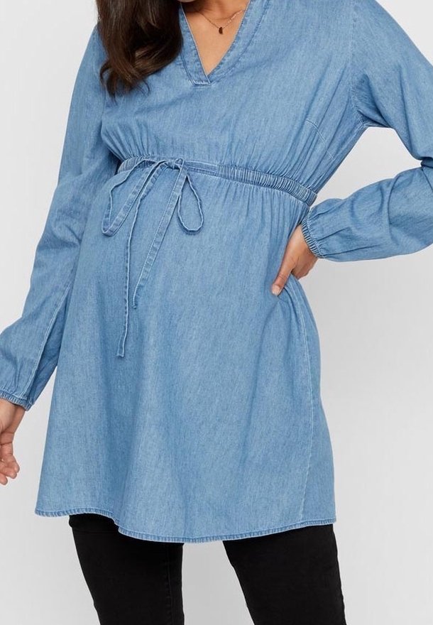 Femme portant une tunique de maternité en denim bleu clair à manches longues avec taille coulissante, associée à un pantalon noir.