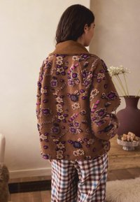 Brązowa bluza polarowa z kolorowym haftem kwiatowym; ma ribbowany kołnierz, luźny krój i jest zestawiona z w kratę spodniami.