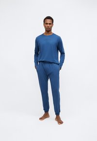 Blå langærmet top og matchende bukser lavet af blødt stof med elastiske manchetter. Har et broderet logo på brystet.