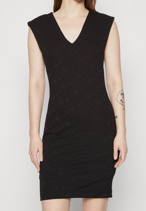 Femme portant une robe noire texturée sans manches avec un décolleté en V, dévoilant un tatouage sur son avant-bras gauche sur un fond uni.