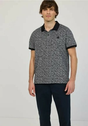 Uomo con polo grigia a maniche corte con colletto nero e motivo a foglie, abbinata a pantaloni scuri, in piedi davanti a uno sfondo neutro.