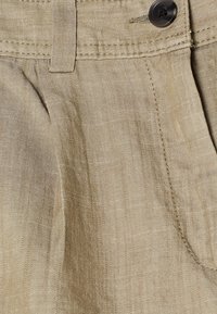 Detailní záběr béžových lněných kalhot s černým knoflíkem, poutkem na pásek, předním skladem a viditelnými švy.