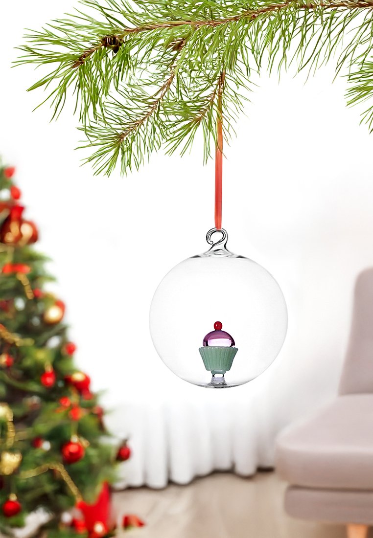 Przezroczysta szklana ozdoba zawierająca wzór babeczki w środku, z purpurową kopułą, czerwoną wisienką i zieloną podstawą, zawieszona na czerwonej wstążce na gałęzi sosny.