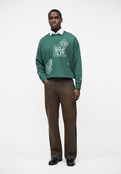 Άνδρας στέκεται φορώντας πράσινο φούτερ με λευκά γραφικά, λευκό πουκάμισο με γιακά, καφέ παντελόνι και μαύρα παπούτσια, μπροστά σε απλό φόντο.