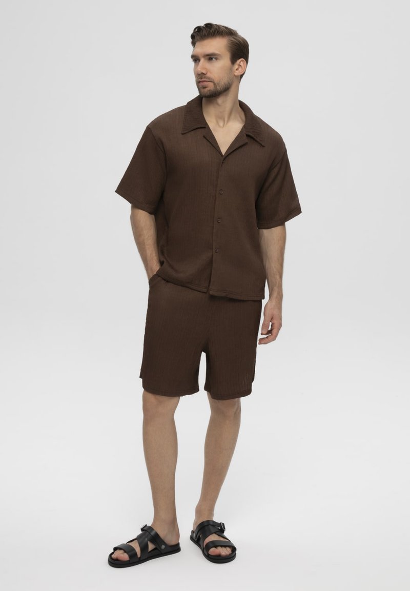 Man draagt een bruin kort mouw knoopshirt en bijpassende korte broek met zwarte sandalen, staand met één hand in de zak tegen een effen achtergrond.