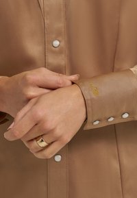 Brązowa satynowa koszula z srebrnymi zatrzaskami, haftowane żółte inicjały na mankiecie, ukazująca gładką teksturę i dopasowany fason. Ręce złożone.