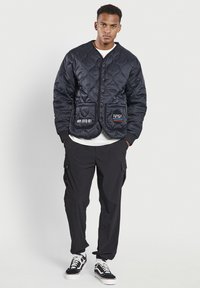 Chaqueta acolchada negra con cuello redondo, botones de presión y dos bolsillos delanteros. Presenta el logo de la NASA y texto en el bolsillo izquierdo, llevada sobre una camisa blanca.