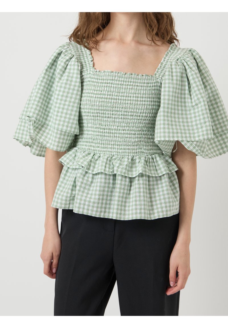 Femme portant un chemisier smocké à carreaux verts et blancs avec des manches bouffantes et un ourlet à volants, associé à un pantalon noir.