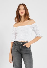 Biely crop top bez ramienok s textúrovaným smockovaním a volánovými rukávmi, v kombinácii s vysokomodrými tmavosivými džínsami. Hladká tkanina a priliehavý strih.