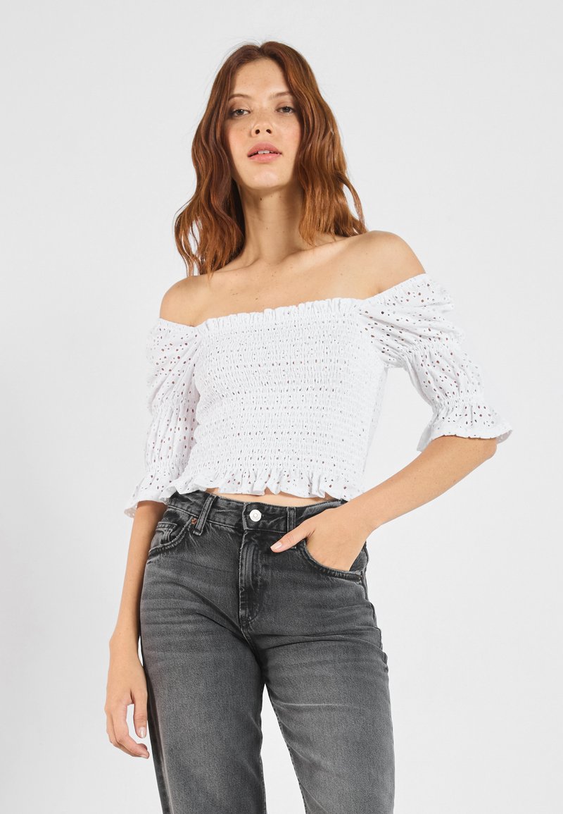 Biely crop top bez ramienok s textúrovaným smockovaním a volánovými rukávmi, v kombinácii s vysokomodrými tmavosivými džínsami. Hladká tkanina a priliehavý strih.