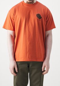 Hombre llevando una camiseta de manga corta naranja con un pequeño gráfico negro y texto en el lado izquierdo del pecho, combinada con pantalones verde oscuro.