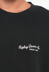 Melna džemperis ar ribotu apkakli, uz kura krūtīm ir balta izšūta teksts: "Replay Denim Co. Kopš 1981. gada". Mīksta auduma tekstūra.
