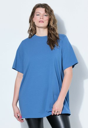 Młoda kobieta z falującymi włosami nosi oversize niebieską koszulkę i czarne skórzane legginsy, stoi na tle jednolitej białej ściany.