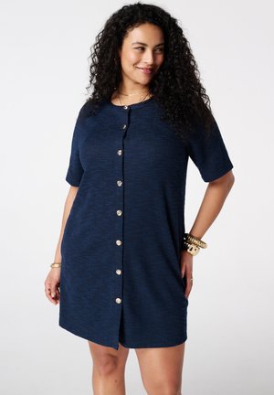 Femme aux cheveux bouclés portant une robe chemise bleu marine à manches courtes, souriante et posant les mains dans les poches avec des bracelets dorés.