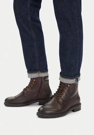 Ciemnobrązowe skórzane botki z sznurowadłami i bocznym zamkiem, noszone z podwiniętymi ciemnoniebieskimi dżinsami.