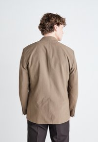 Brauner strukturierter Blazer mit Reverskragen, mittigem Schlitz und langen Ärmeln, getragen über dunkleren Hosen, von hinten betrachtet.
