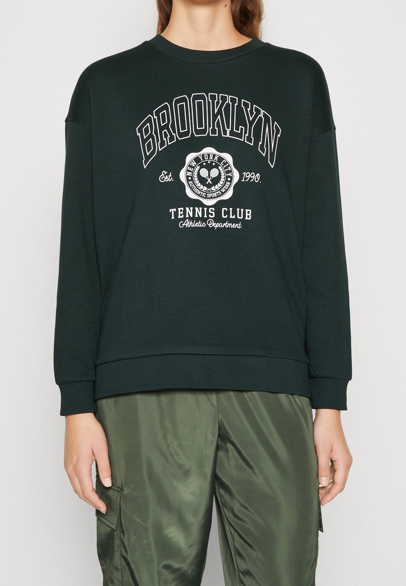 Personne portant un sweat-shirt vert foncé avec le texte et le logo « Brooklyn Tennis Club New York City », associé à un pantalon cargo vert olive.