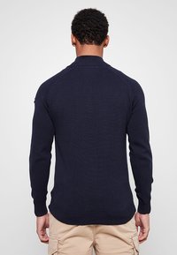 Granatowy sweter z dzianiny z długimi rękawami i prążkowanym kołnierzem. Posiada bezszwowe ramiona i prosty dół u pasa. Miękka tekstura.