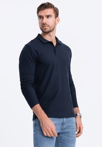 Mörkblå långärmad polo med klassisk krage, tillverkad av mjukt tyg, med en subtil textur och en ren, figursydd design.