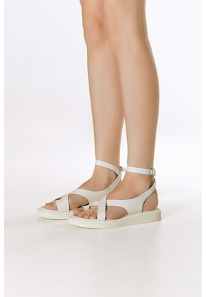 Nogi w białych płaskich sandałach z paskami na kostce i wycięciami na palce na jednolitym białym tle.