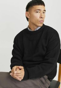 Melnas adītas džemperis ar tekstūras kvadrātiem, ar apaļu kakla izgriezumu un ribotiem piedurkņu galiem. Apvienots ar apakšā valkātu krekliņu ar uzvalka apkakli un svītrām.