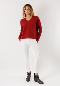 Maglione rosso a maglia con scollo a V, orlo a coste, abbinato a pantaloni bianchi aderenti e stivaletti neri alla caviglia. Design semplice, texture liscia.