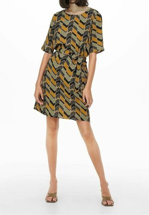 Femme portant une robe longueur genoux avec un motif géométrique chevron en moutarde, olive, noir et beige, associée à des sandales à bouts ouverts.
