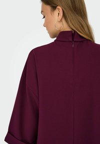 Abito borgogna con un colletto alto, maniche corte e una cerniera nascosta sul retro. Tessuto liscio con un design pulito e senza ornamenti.