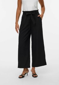 Zwarte wijde broek met een hoge taille, voorzien van een stoffen riem, plooien aan de voorkant en een gladde textuur, gecombineerd met zwarte hakken sandalen.