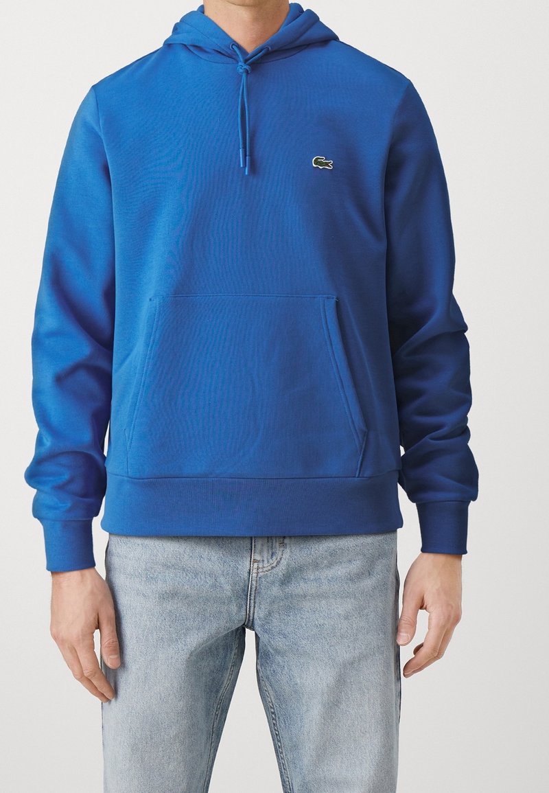 Homme portant un sweat à capuche bleu vif avec poche avant et petit logo crocodile, associé à un jean bleu clair, debout devant un fond uni.