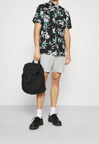 Camisa negra de manga corta con estampado floral en blanco y verde, pantalones cortos grises, mochila negra con asa superior y zapatos negros con calcetines blancos.