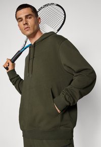 Sweatshirt verde-oliva feito de tecido macio, com um bolso frontal, capuz ajustável com cordão e punhos canelados. Raquete de ténis segurada ao lado.