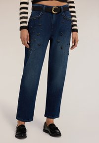 Jeans de mezclilla azul oscuro con un ajuste holgado, adornados con acentos de cuentas negras en la parte frontal, y cuenta con un cinturón negro con una hebilla dorada.