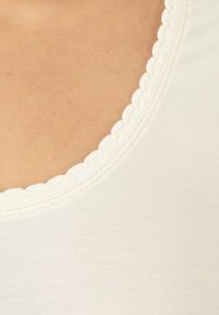 Top bianco in cotone con scollatura a onde. Il tessuto ha una consistenza liscia con sottili dettagli di cucitura lungo i bordi.