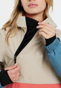Beige Jacke mit pinken und blauen Akzenten; hat einen Reißverschluss vorne, elastische Bündchen und Logodetails. Glatte Textur, sportliches Design.