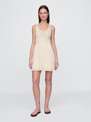 Young woman with straight brown hair wearing a sleeveless cream knit dress and white flip-flops, standing against a plain light background.