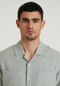 Világos zöld, rövid ujjú ing, könnyű anyagból készült, tábori gallérral, két mellzsebbel és fehér gombos záródással.