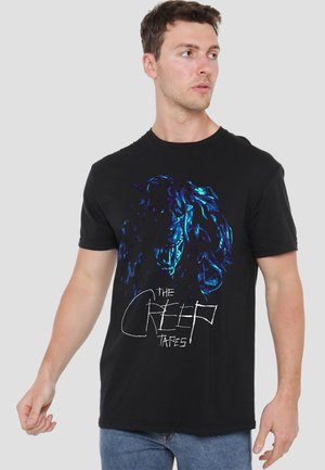 Man wearing black T-shirt with blue neon Medusa head graphic and "The Creep Tapes" text, standing against plain gray background.