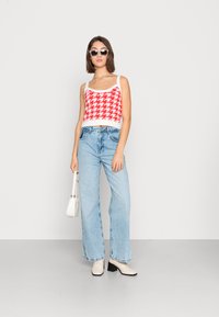 Top crop tricotado em padrão pied-de-poule vermelho e branco, jeans de perna larga em azul claro e botas de salto bege. Acessórios incluem óculos de sol redondos e uma bolsa branca.